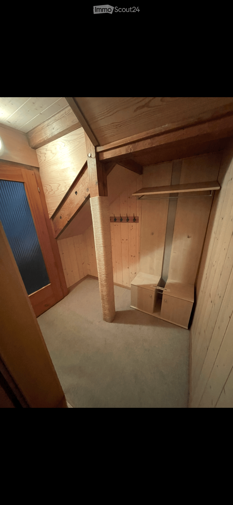 An unfurnished room with wooden walls, a small window, a built-in shelf, and a coat rack. The room has a slanted ceiling and a carpeted floor.