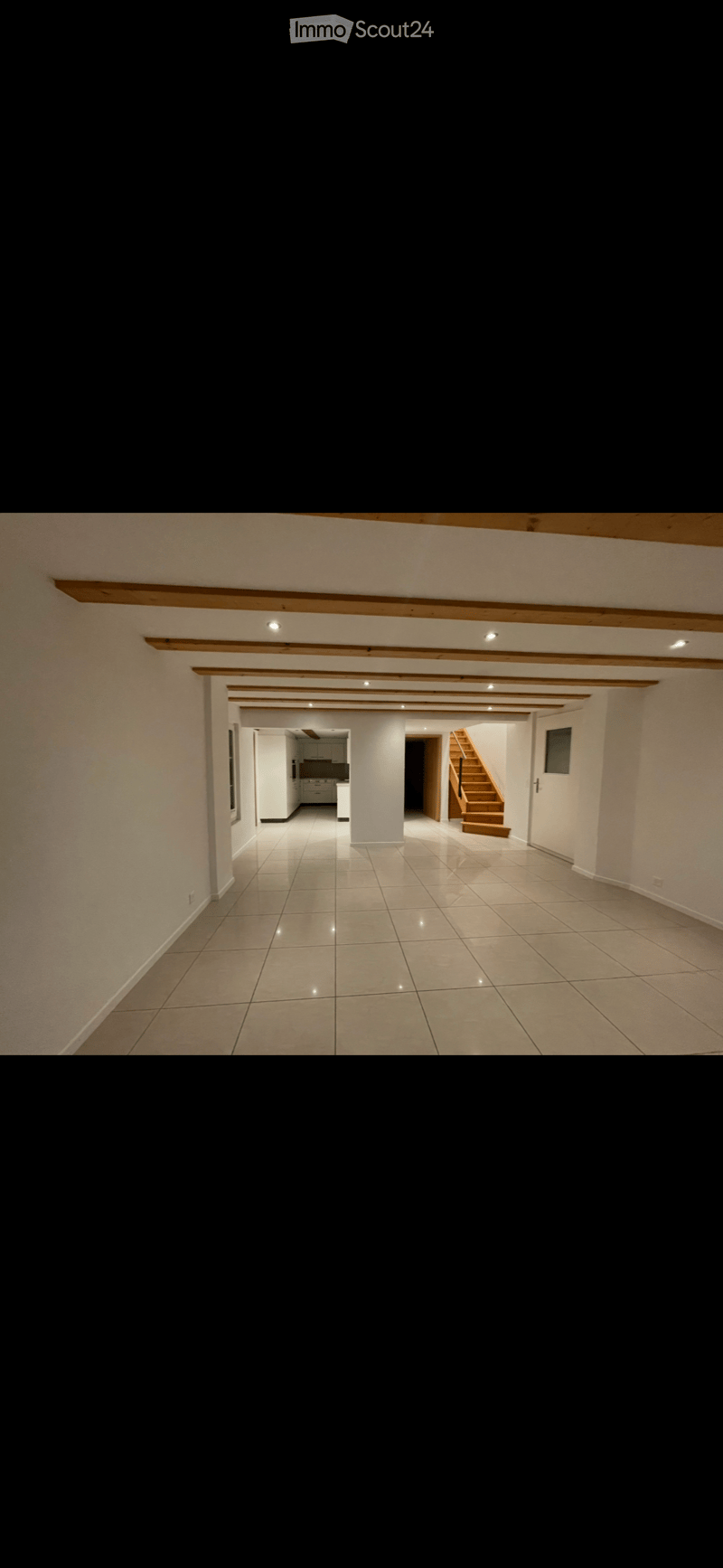 Empty hallway with tiled floor, white walls, and wooden beams on the ceiling. Visible doorways lead to a kitchen and a staircase.
