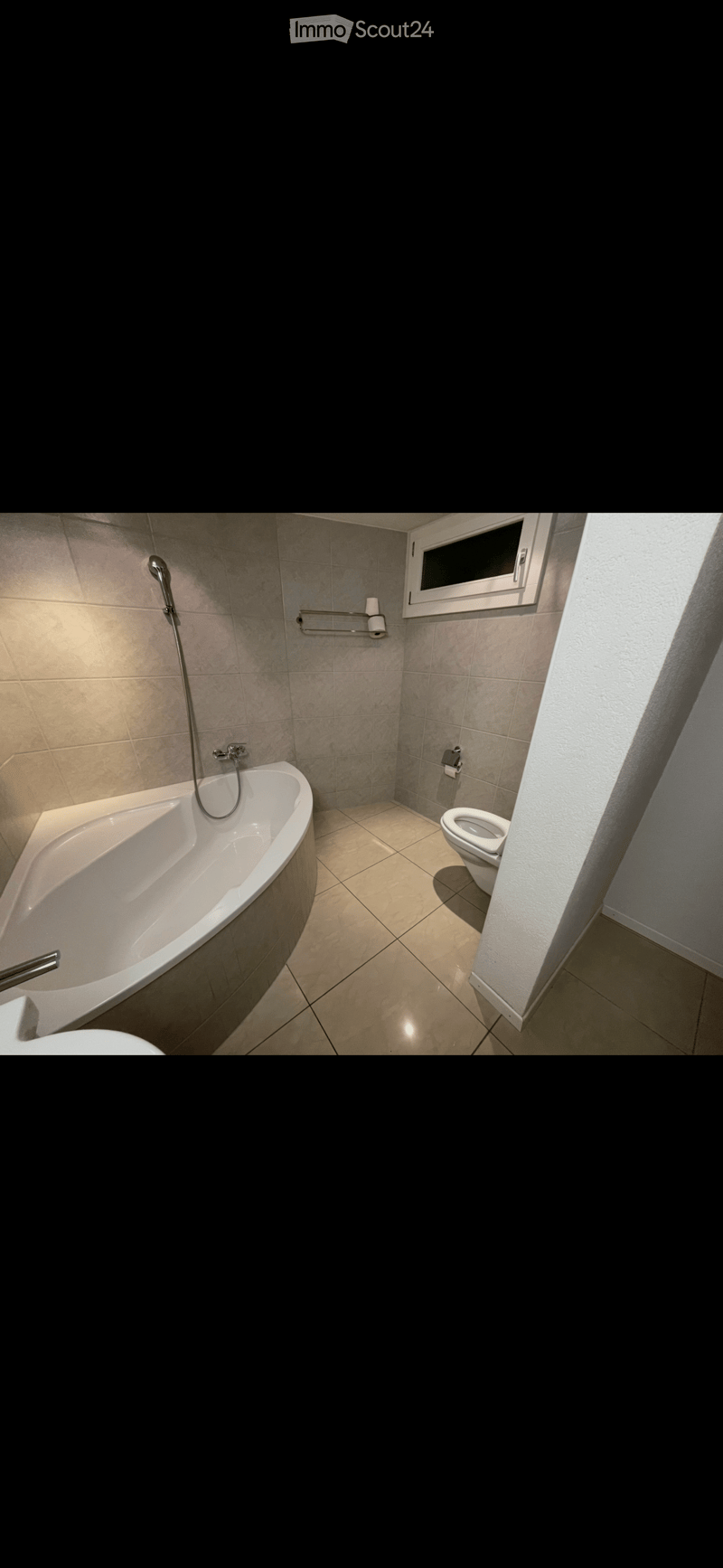 Modern bathroom with a bathtub, toilet, showerhead, and towel rack. Light-colored tiles and a window provide natural light.