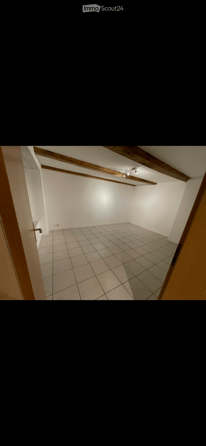 Empty room with tiled floor, white walls, wooden beams on the ceiling, and a single light fixture.
