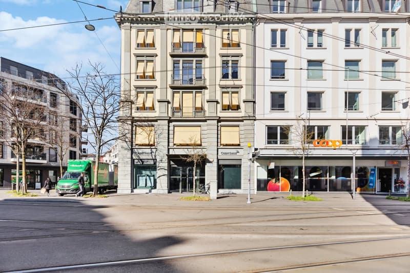 A multi-story building with large windows and balconies, featuring commercial storefronts at the ground level, including a 'Coop' supermarket and a 'Credit Suisse' bank branch. The building has a classic architectural style with ornate detailing and is si