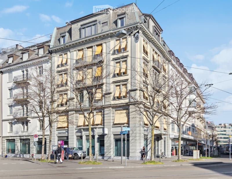 A multi-story building with classic architecture, large windows, and balconies. The facade is light-colored with decorative elements. The ground floor has commercial storefronts, and there are trees and street signs visible in the foreground.