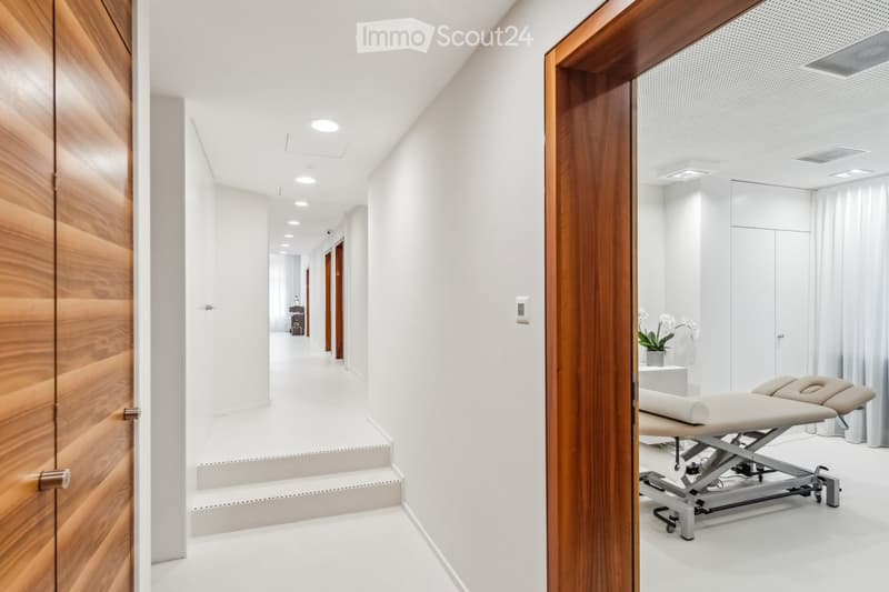 Modern, clean hallway with white walls, recessed lighting, and wooden paneling on one side. A room visible at the end contains a massage table and a small plant on a stand.