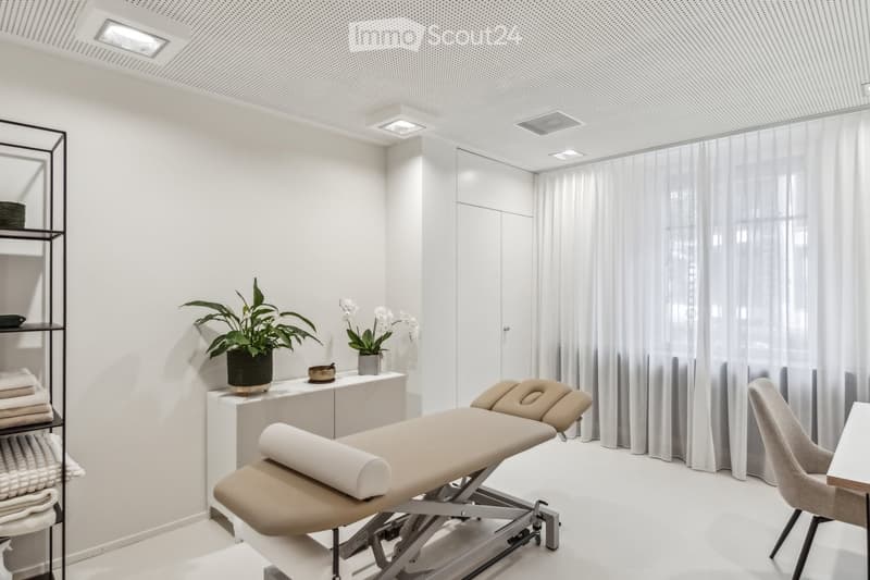 Modern, minimalist room with a massage table, a small table with a plant and decorative items, a chair, and a shelf with towels.