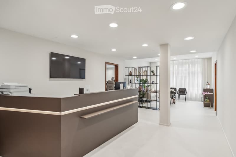 Modern reception area with a sleek reception desk, a large wall-mounted screen, a few chairs, small tables, and decorative plants.
