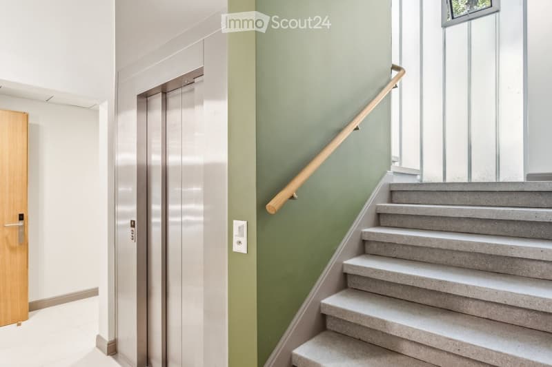 Modern hallway with a staircase featuring grey concrete steps and a wooden handrail, adjacent to an elevator and a wooden door.
