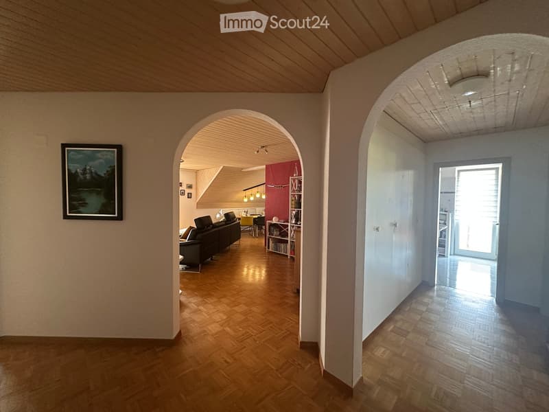 Spacious hallway with wooden flooring, arched doorways leading to a living area and another room, and a framed picture on the wall.