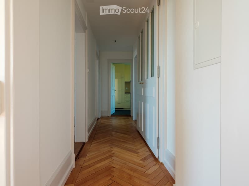 A long, narrow hallway with wooden herringbone flooring, white walls, and several doors leading to other rooms.