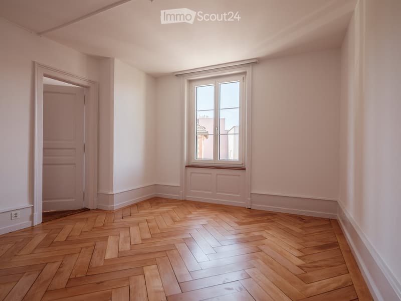 Empty room with light pink walls, wooden herringbone flooring, and a large window with white frame and blinds. There is a doorway leading to another room.