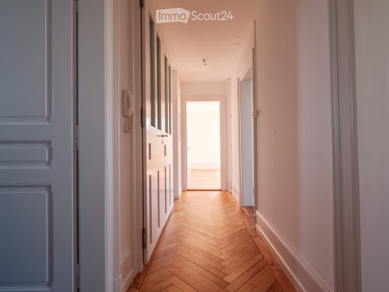A narrow hallway with wooden herringbone flooring, light-colored walls, and white doors on both sides. Natural light streams in from an open doorway at the end.