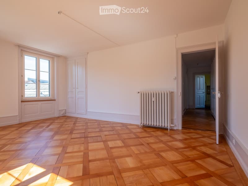 Empty room with wooden herringbone flooring, white walls, a radiator, and a window with a view of rooftops.
