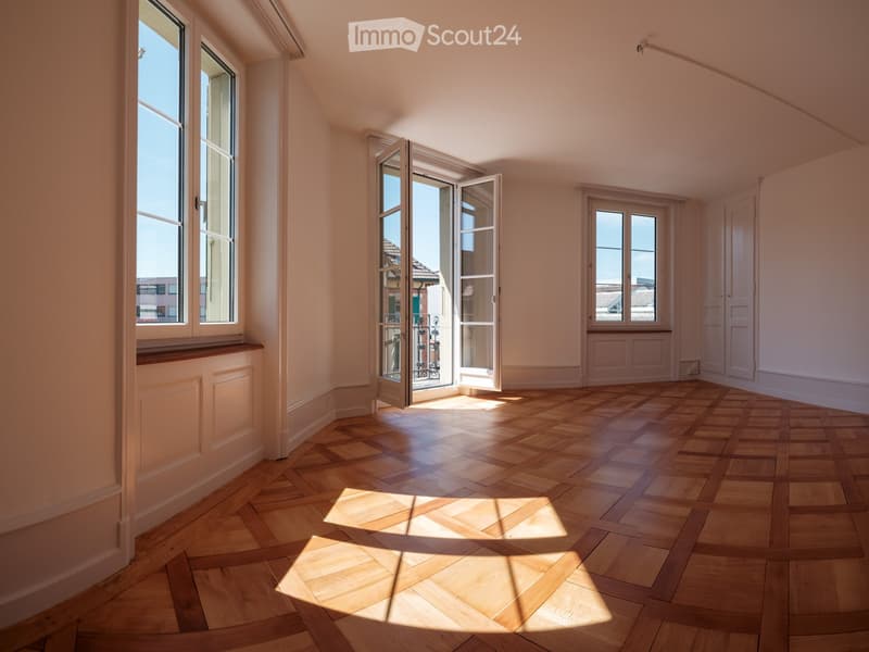 Empty room with wooden herringbone flooring, large windows, and sliding glass doors leading to a balcony.
