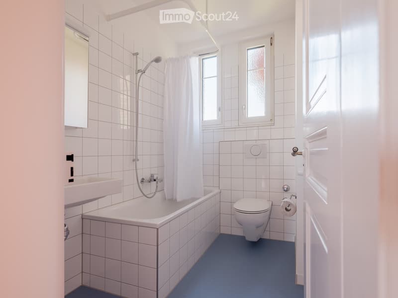 Modern bathroom with white tiled walls, a bathtub, toilet, sink, and a shower with a curtain. Blue flooring and two windows provide natural light.