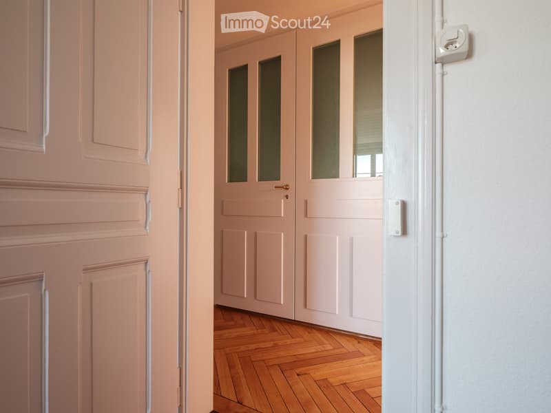 A narrow hallway with light pink walls, wooden flooring, and double doors with frosted glass panels leading to another room.