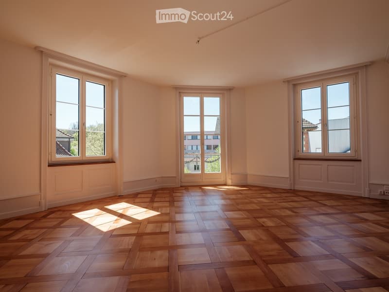 Empty room with wooden herringbone flooring, large windows, and a door leading outside. Light-colored walls and baseboards.
