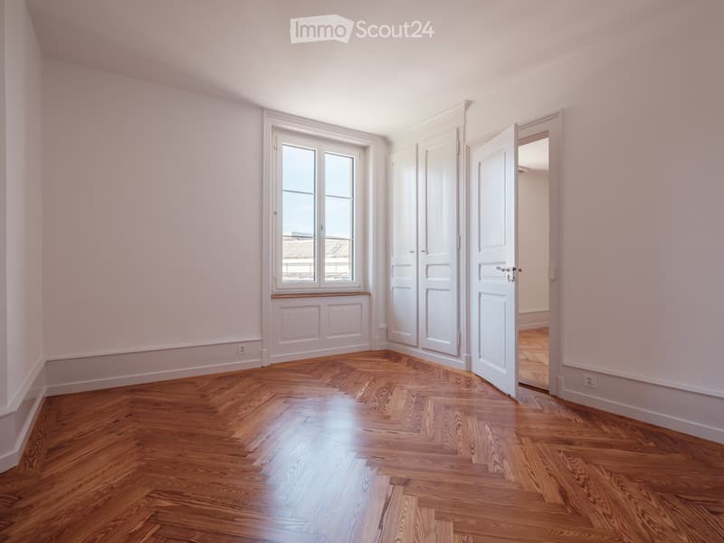 Empty room with wooden herringbone flooring, white walls, and large windows with white frames. The room features built-in cabinetry and an open doorway leading to another space.