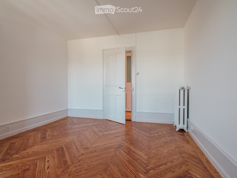Empty room with wooden herringbone flooring, white walls, and a radiator on one side. An open door leads to another room.