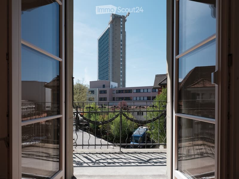 Balcony with metal railing, overlooking a cityscape with a tall building and surrounding structures.