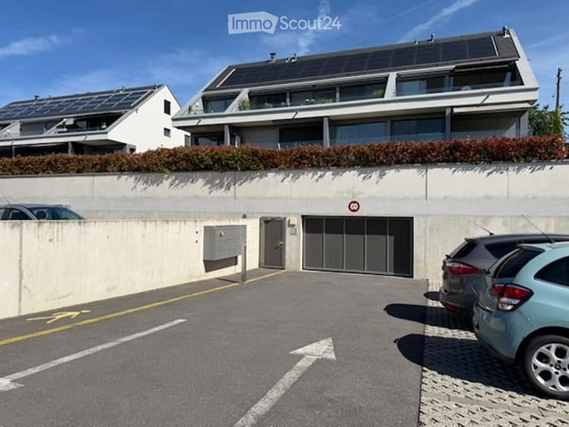 Modern multi-story building with solar panels on the roof, garage entrance, and adjacent parking area with several cars.