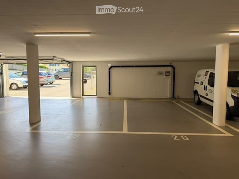 Underground parking area with marked parking spaces, concrete pillars, and a few parked cars visible through the open entrance.