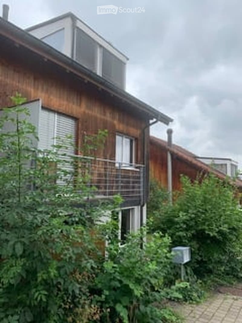 Two-story wooden building with a flat roof and balcony, surrounded by greenery.