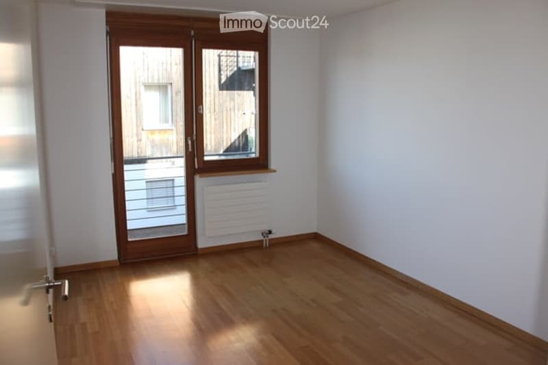 Empty room with wooden flooring, white walls, large window, and sliding glass door leading outside.