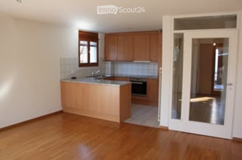 Bright, unfurnished kitchen with wooden flooring, white walls, and a kitchen island with a sink and oven. The space is clean and well-lit with natural light from a window.