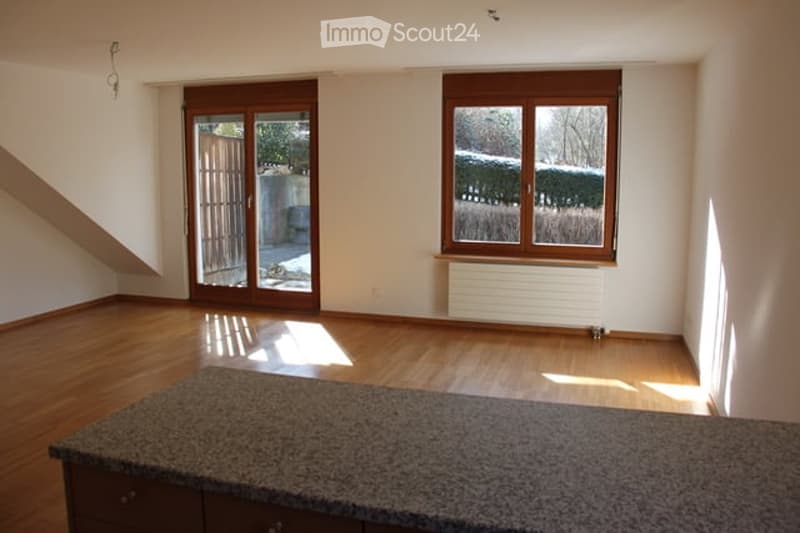 Empty room with wooden flooring, large windows, sliding glass door leading to outdoor area, and a granite countertop with cabinets.