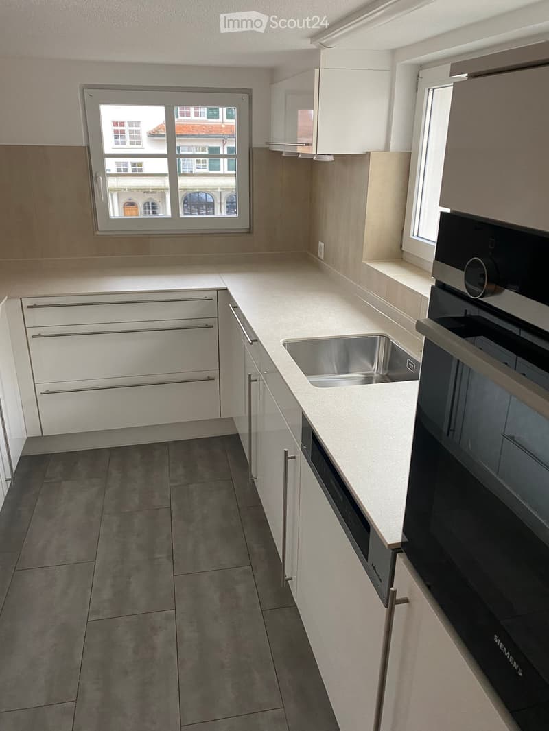 Modern kitchen with white cabinets, stainless steel sink, and built-in oven. Features large windows with city view.