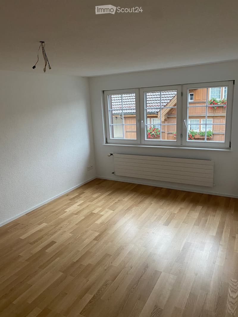 Empty room with light wooden flooring, white walls, and large windows showing a view of a neighboring building with flower boxes.