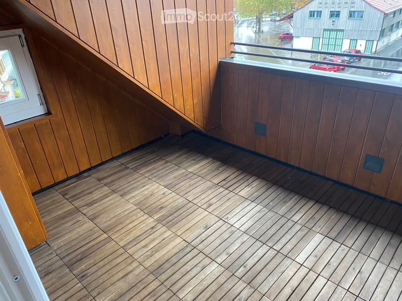 Balcony with wooden decking, wooden panel walls, and a metal railing overlooking a parking area with several cars.