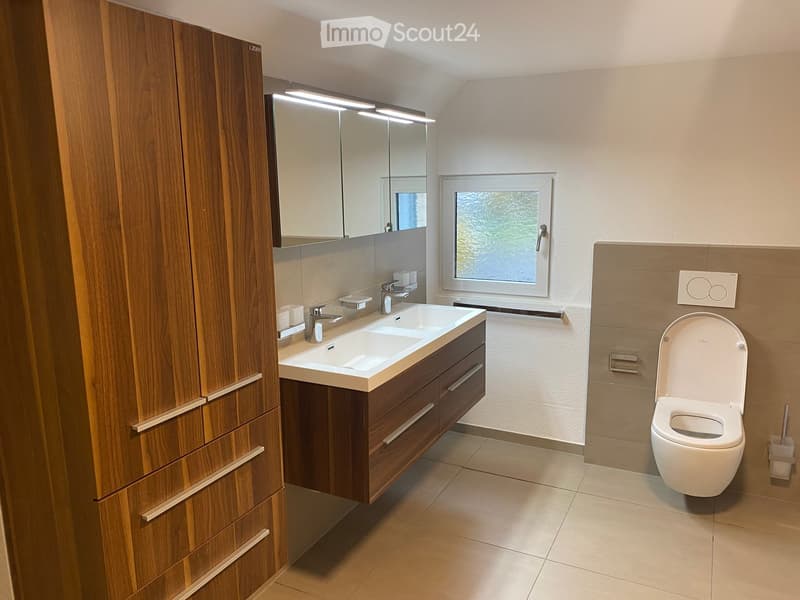 Modern bathroom with a double sink, wooden cabinet, large mirror, and toilet. Features a window with a view of greenery.