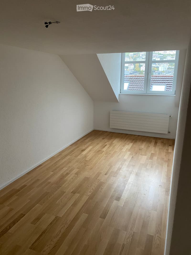 Empty room with wooden flooring, white walls, and a large window offering a view of rooftops. A radiator is mounted on the wall below the window.