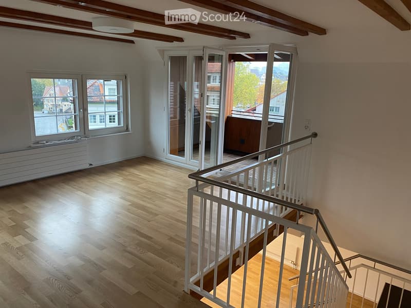 Empty room with wooden flooring, white walls, large windows, and a staircase leading to a balcony area.