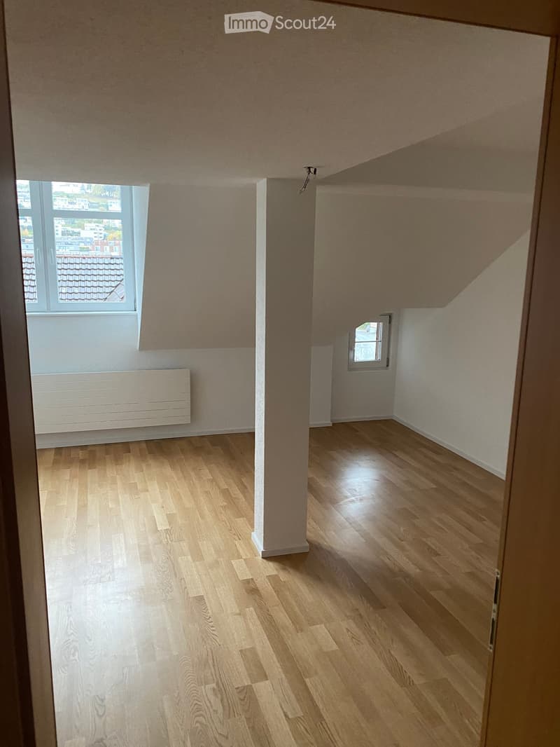 Empty attic room with wooden flooring, slanted ceilings, and two windows offering city views.