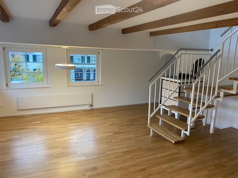 Empty room with wooden flooring, white walls, exposed wooden beams on the ceiling, two windows, a radiator, and a staircase leading upwards.