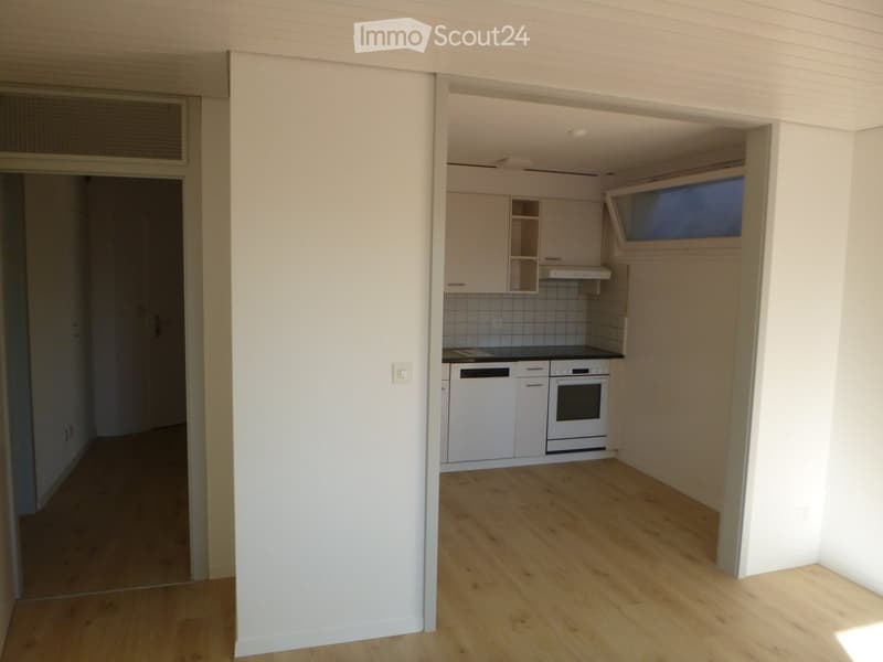 Empty kitchen with white cabinets, a black countertop, and a small window above the sink. The floor is light wooden, and the walls are white.