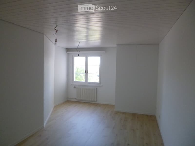 Empty room with white walls, wooden flooring, and a single window with white frame. Exposed electrical wiring and a radiator are visible.