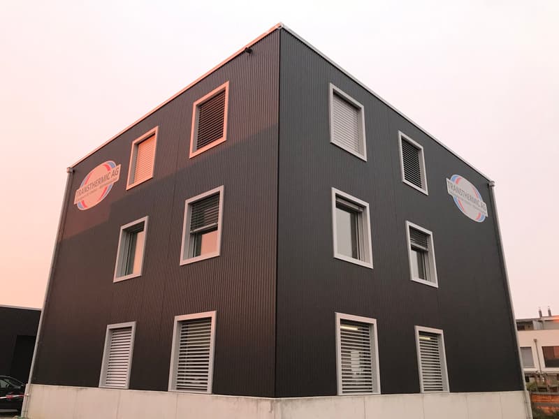 Modern two-story building with dark brown metal siding, multiple windows with white frames, and company logos on the facade.