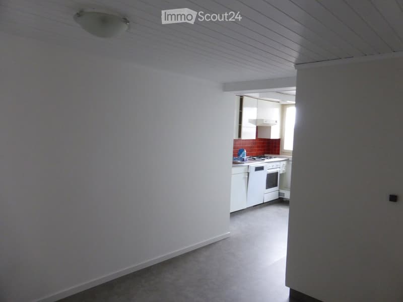 Open-plan living space with a small kitchen area featuring white cabinets, a stove, oven, and dishwasher. The main area has white walls and a carpeted floor.