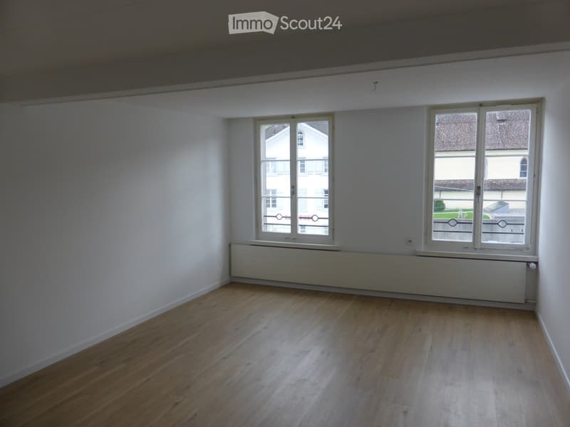 Empty room with wooden flooring, white walls, and two large windows offering views of neighboring buildings.
