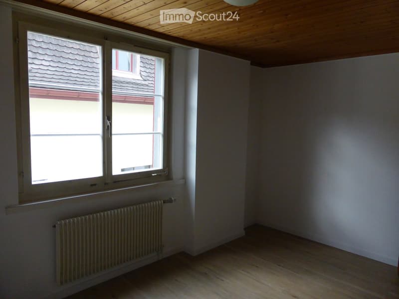 Empty room with wooden flooring, white walls, large windows, and a radiator under the windows.