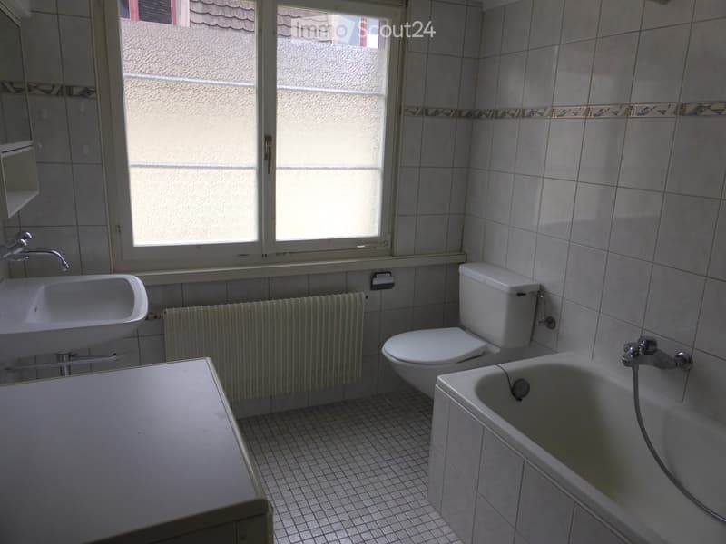 Bathroom with white tiles, a sink, toilet, bathtub, radiator, and a mirror. The room has a window with frosted glass and a decorative tile border near the top of the walls.