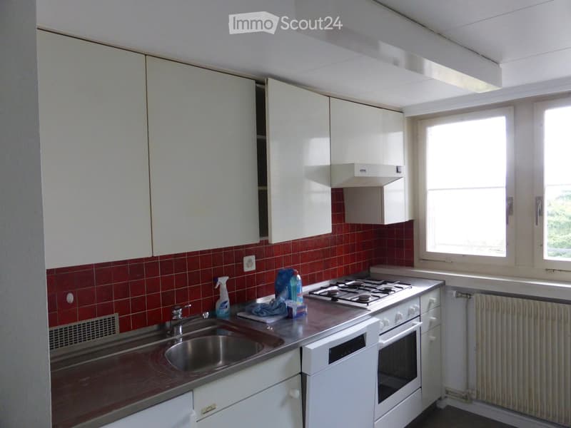 A compact kitchen with white cabinets, a stainless steel sink, a gas stove, and an oven. The walls are tiled in red, and there is a window providing natural light.