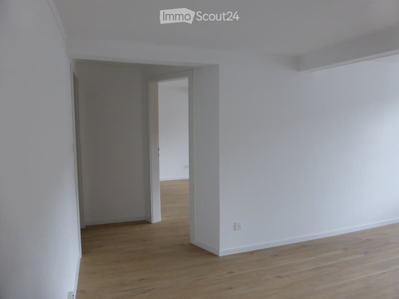 Empty room with white walls and wooden flooring, featuring an open doorway leading to another room.