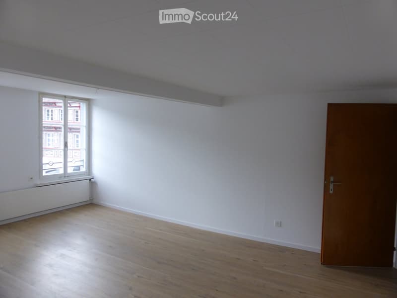 Empty room with white walls, wooden floor, and a single window with a view of a building outside. There is a closed wooden door on the right side.