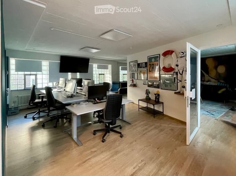 Modern office space with light wooden flooring, large windows, desks with chairs, multiple screens, and decorative items including a small table with figurines and artwork on the wall.