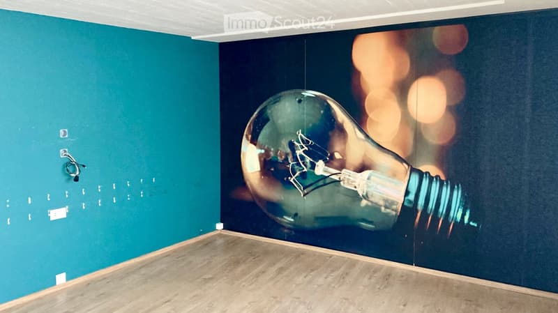 Empty room with wooden flooring and two walls, one teal and one dark blue, featuring a large light bulb mural on the dark blue wall.