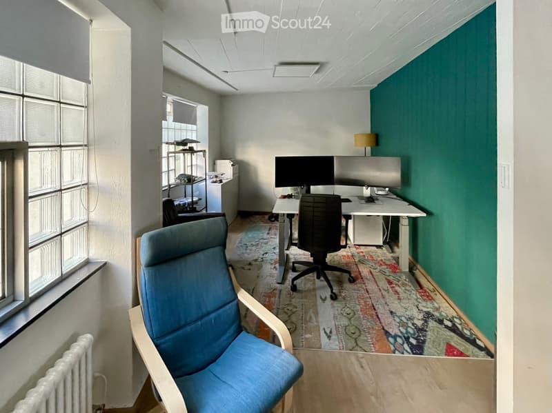 Office space with a blue armchair, desk, screen, lamp, and table. The room features large windows, a patterned rug, and a green accent wall.