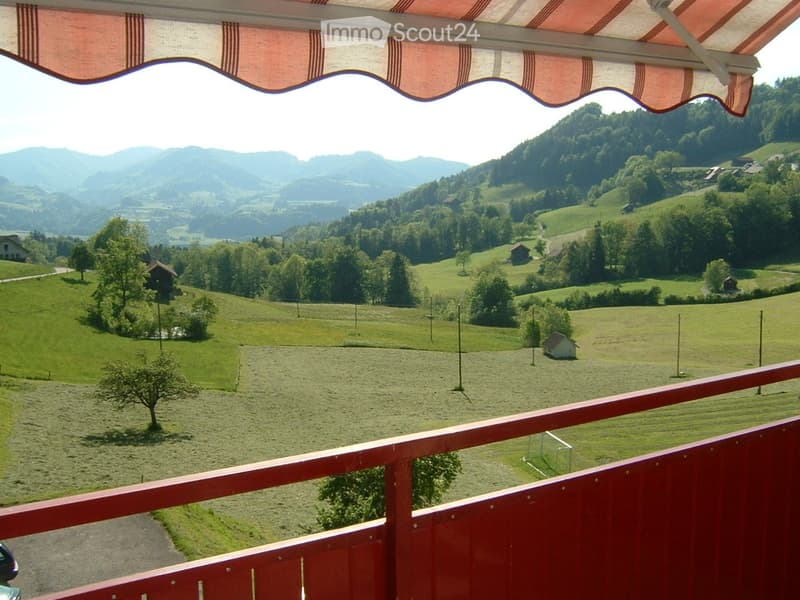 Balcony with red wooden railing, overlooking a scenic valley with green fields, scattered trees, and distant mountains.